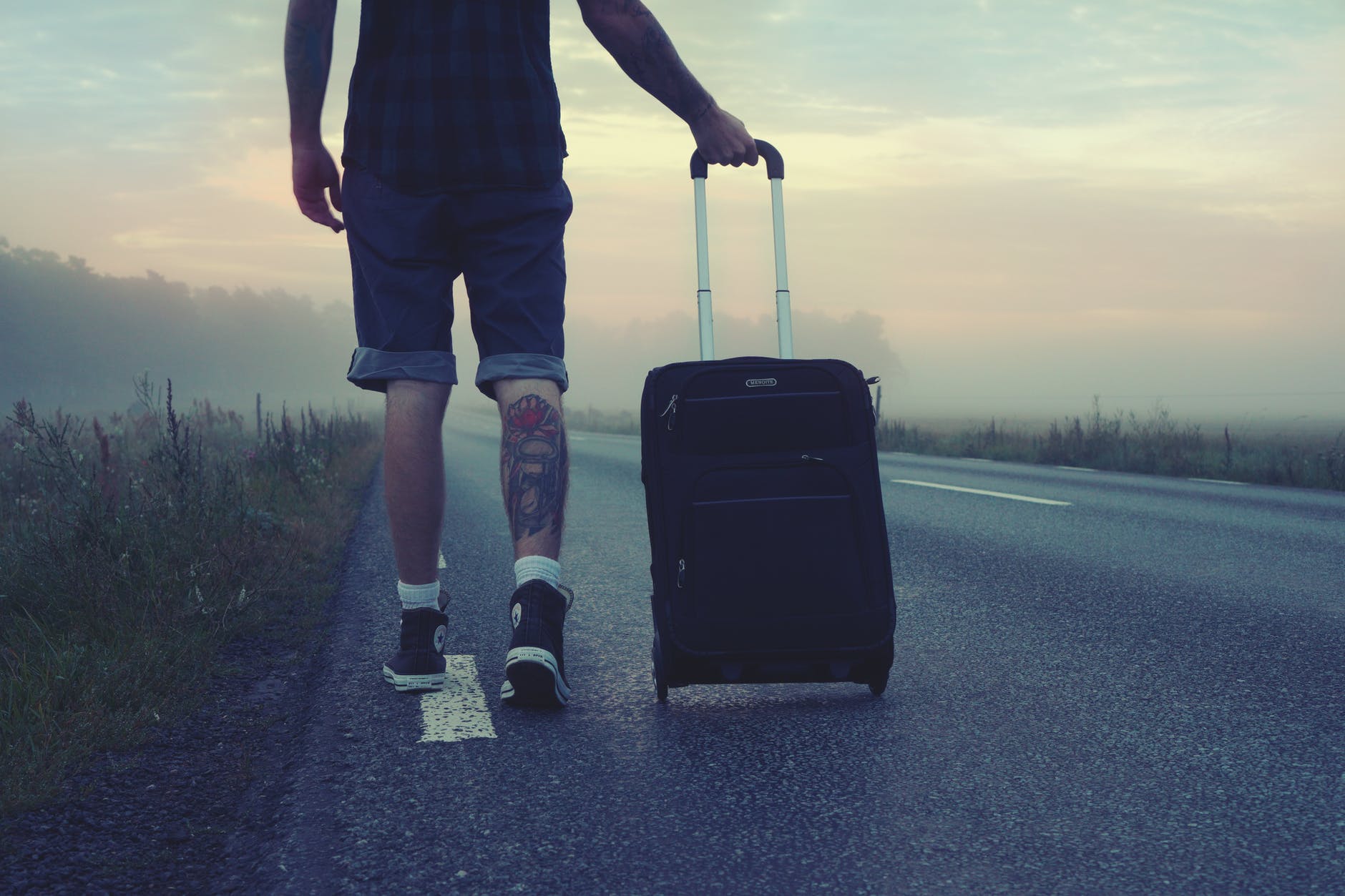 man with leg tattoo rolling suitcase down a long road